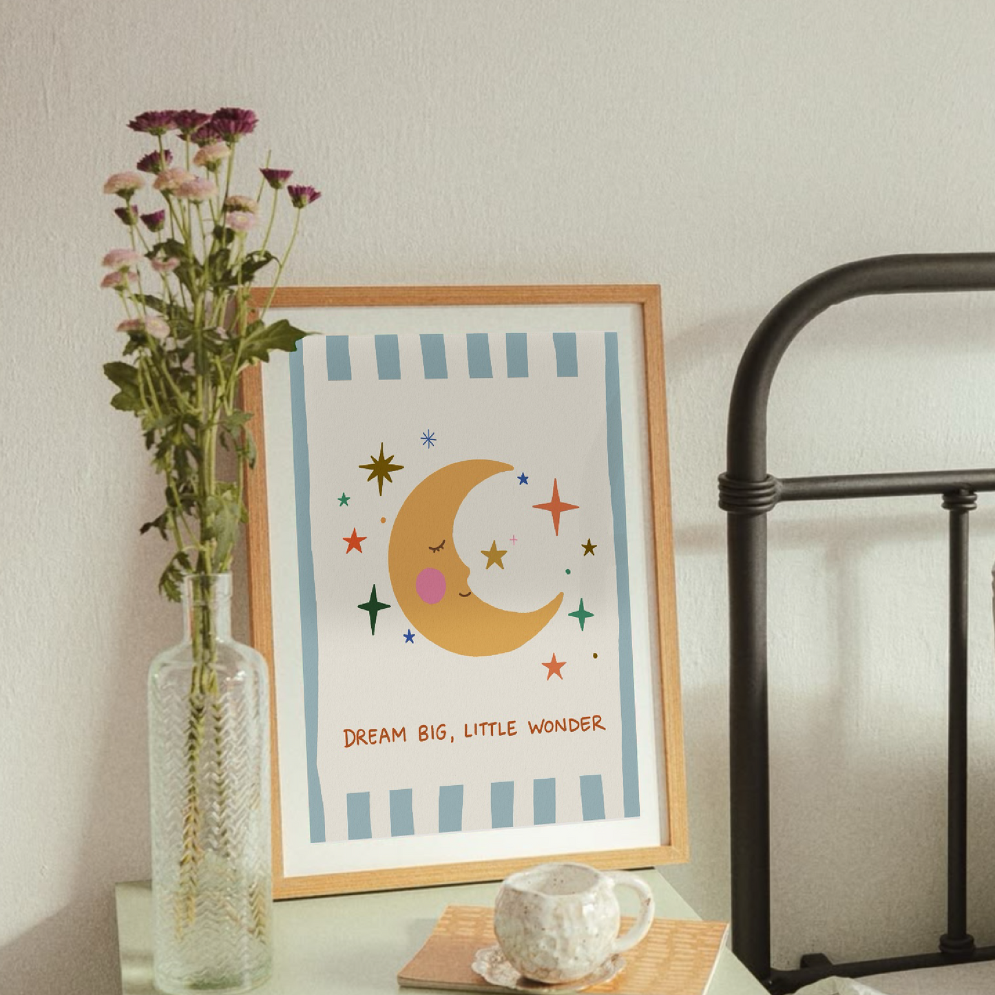 Konsttryck med måne och stjärnor i träram, placerad i en inspirerande miljö med en vas blommor och en kaffekopp.