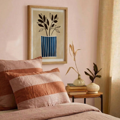 Minimalistisk väggkonst med blårandig vas – Inramad poster i träram hängandes på en rosa vägg i ett harmoniskt sovrum. Miljöbilden visar en bäddad säng med mjuka linnekuddar i terracotta och rosa ränder som matchar den botaniska tavlan.