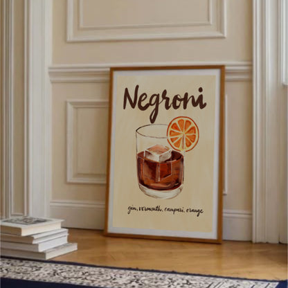 Negroni-poster i ram på golv lutad mot vägg med böcker och vita blommor