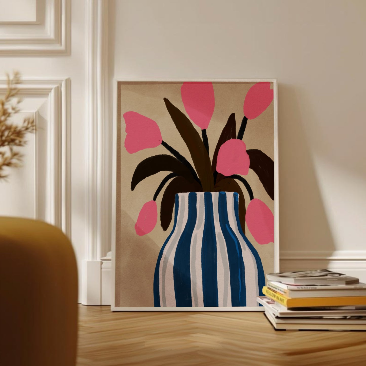 Inramad botanisk illustration med rosa tulpaner i en blå och vit randig kruka. Konstverket står lutad mot väggen i ett vardagsrum med en hög böcker framför.