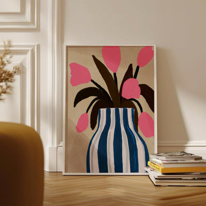 Inramad botanisk illustration med rosa tulpaner i en blå och vit randig kruka. Konstverket står lutad mot väggen i ett vardagsrum med en hög böcker framför.