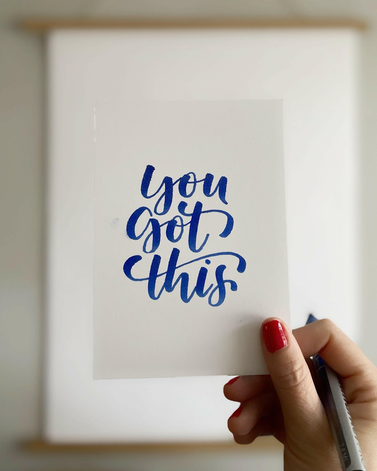 En hand som håller upp ett vitt kort med den handstils-kallograferade texten You got this i blått bläck, skapad med en penselpenna.