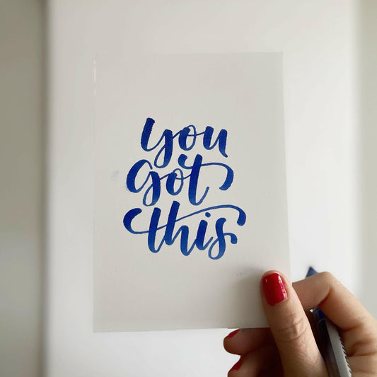 En hand som håller upp ett vitt kort med den handstils-kallograferade texten You got this i blått bläck, skapad med en penselpenna.