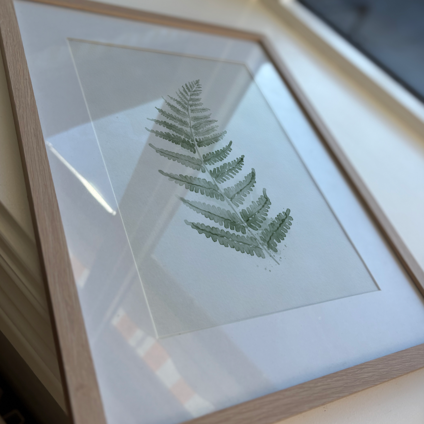 En inramad botanisk poster med gröna ormbunksblad som ligger plant på en yta, visad i en modern och minimalistisk miljö.