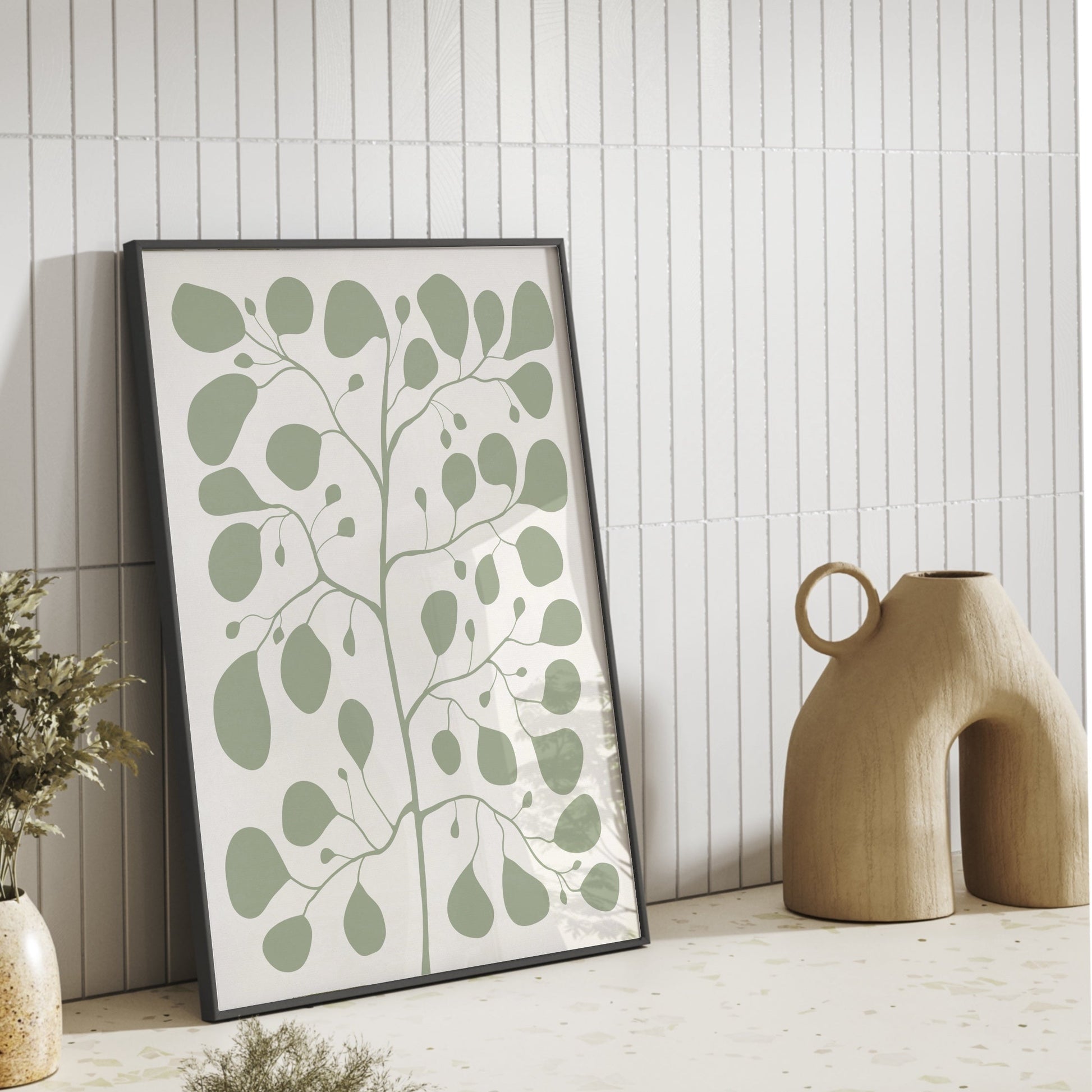 Stilren inredning med en inramad Pilea-poster i färgen jade som lutar mot en vit kakelvägg bredvid en modern beige keramikvas.