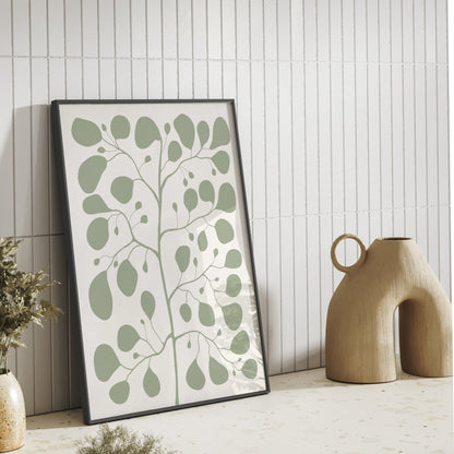 Stilren inredning med en inramad Pilea-poster i färgen jade som lutar mot en vit kakelvägg bredvid en modern beige keramikvas.