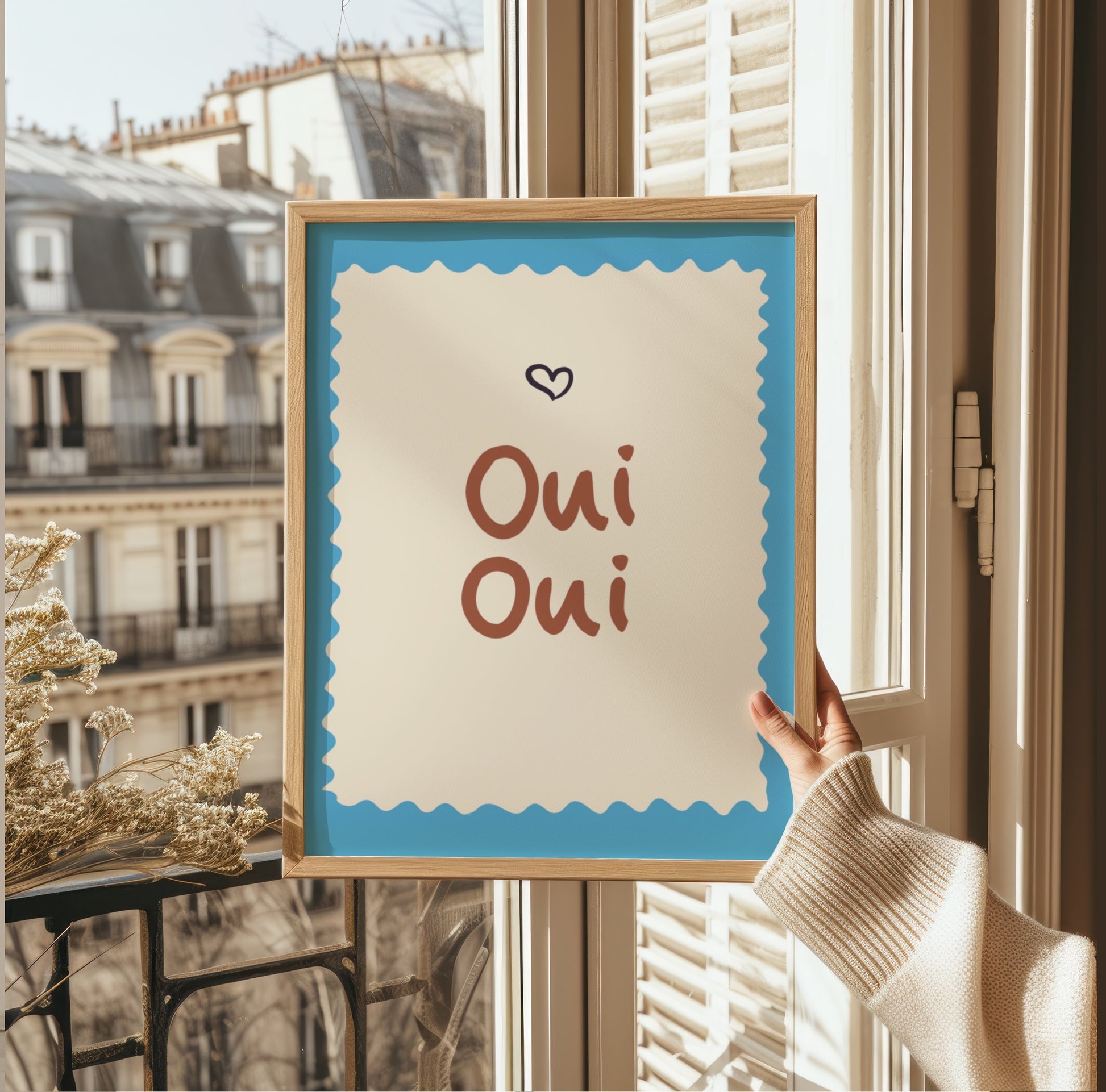 En person håller upp Oui Oui-postern framför ett fönster med utsikt över takåsar i Paris, vilket förstärker den franska känslan.