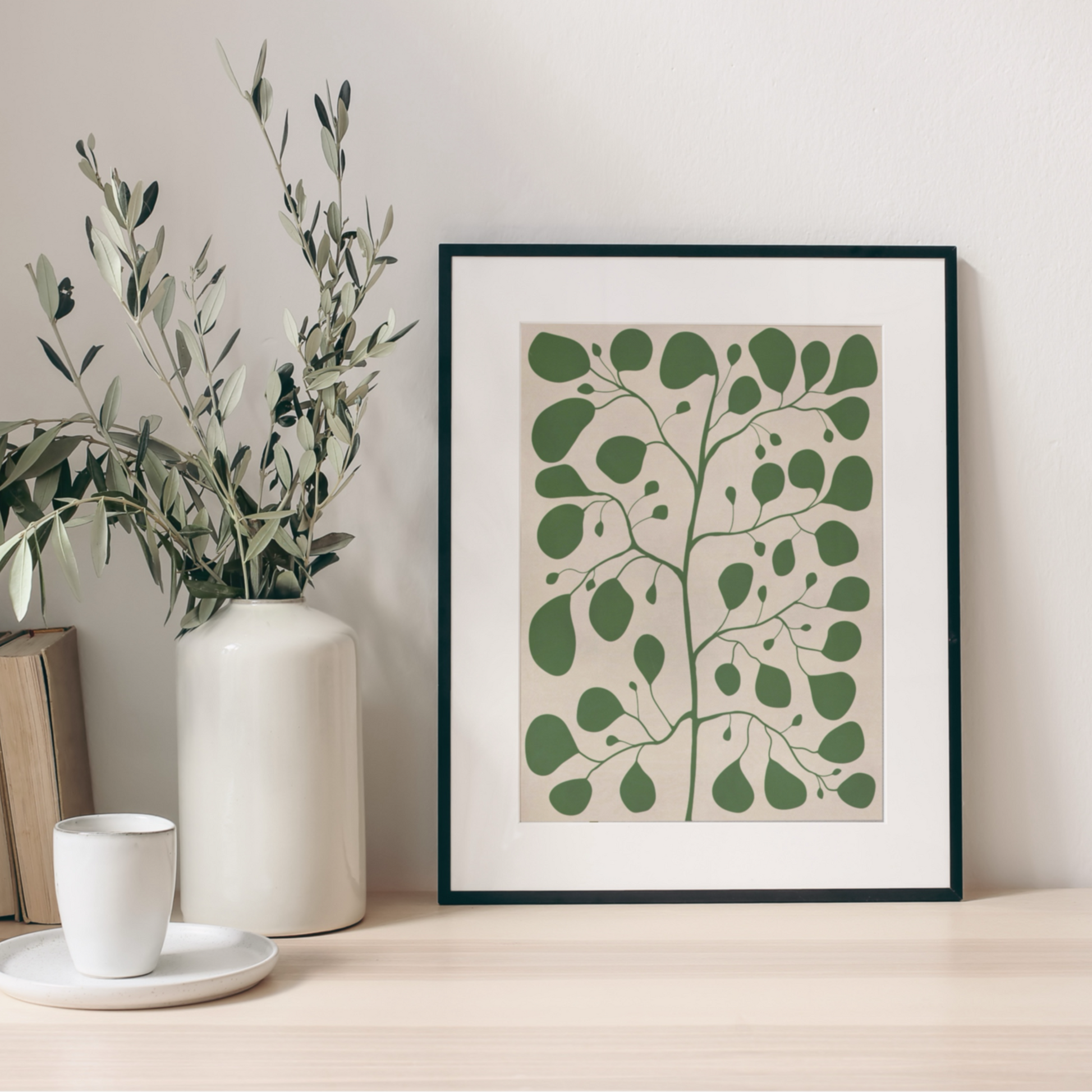 Närbild på Pilea-poster i svart ram på ett ljust träbord bredvid en vit vas med olivkvistar. En dämpad pastellbakgrund som ger en bohemisk underton till inredningen.