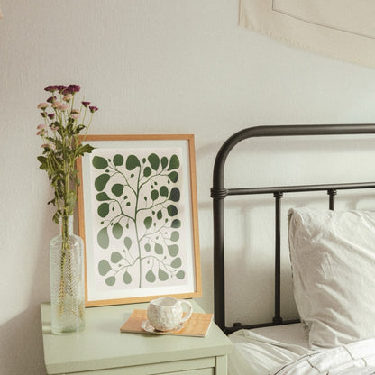 Pilea Jade poster inramad i en ljus träram som står lutad mot väggen på ett grönt nattduksbord i ett harmoniskt sovrum. Intill tavlan syns en vas med lila blommor och en kopp kaffe, vilket visar hur den botaniska illustrationen skapar en lugn och ombonad atmosfär vid sängplatsen.