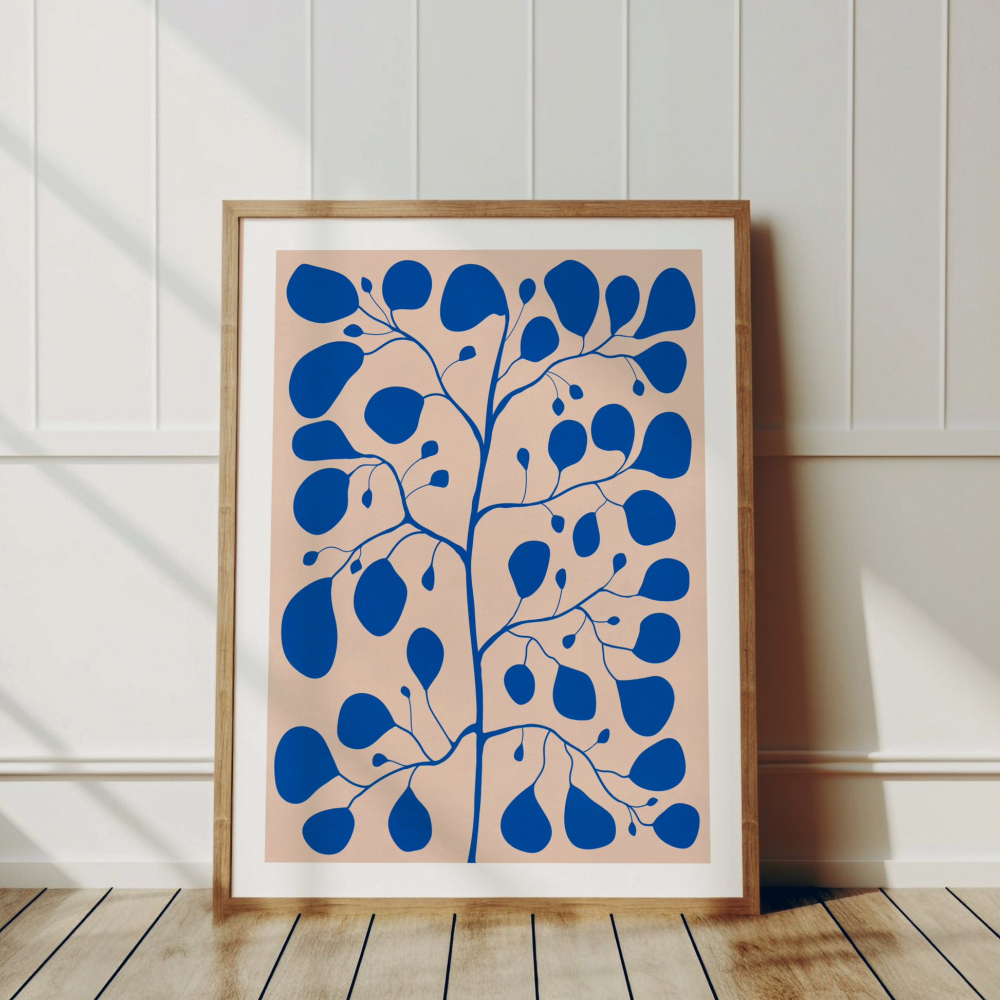 Poster med blå Pilea Peperomioides och vit kant, lutad mot vit vägg – skandinavisk inredningsstil.