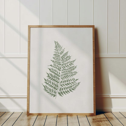 En stor botanisk tavla med gröna ormbunskblad ståeendes i träram lutad mot en vägg.